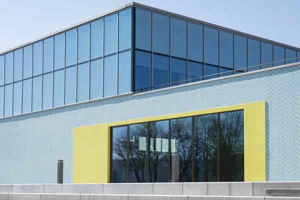 Generous floor-to-ceiling windows bring daylight into the reception area. Bright yellow plaster frames set fresh accents in the otherwise quiet color scheme of the façade.